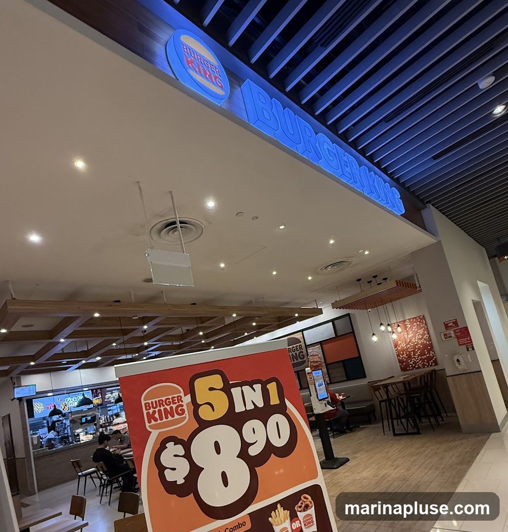 A Burger King promotional sign is in the foreground, advertising a "5 IN 1" deal for $8.90. In the background, the Burger King logo and signage are visible above the restaurant counter. The interior of the fast-food restaurant is also visible, with seating areas and a food preparation area.