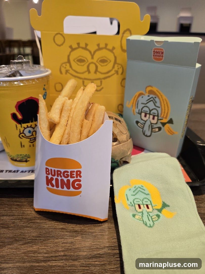 A Burger King french fry container with the Burger King logo is in the foreground. Behind it, a yellow box with Spongebob Squarepants characters and a light blue box with Squidward Tentacles characters are visible. A cup with Spongebob graphics and a pair of light green socks with Squidward Tentacles graphics are also present.