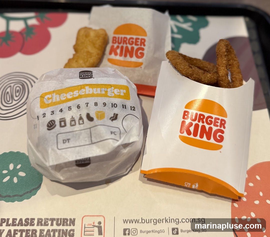 A Burger King cheeseburger, a hash brown, and onion rings are arranged on a tray liner. The burger is wrapped in white paper with the Burger King logo and the word "Cheeseburger" printed on it. The onion rings are in a white paper container with the Burger King logo. The hash brown is in a white paper bag with the Burger King logo. The tray liner has a graphic design with tomatoes and onions, and text that says "PLEASE RETURN TRAY AFTER EATING" and "www.burgerking.com.sg".