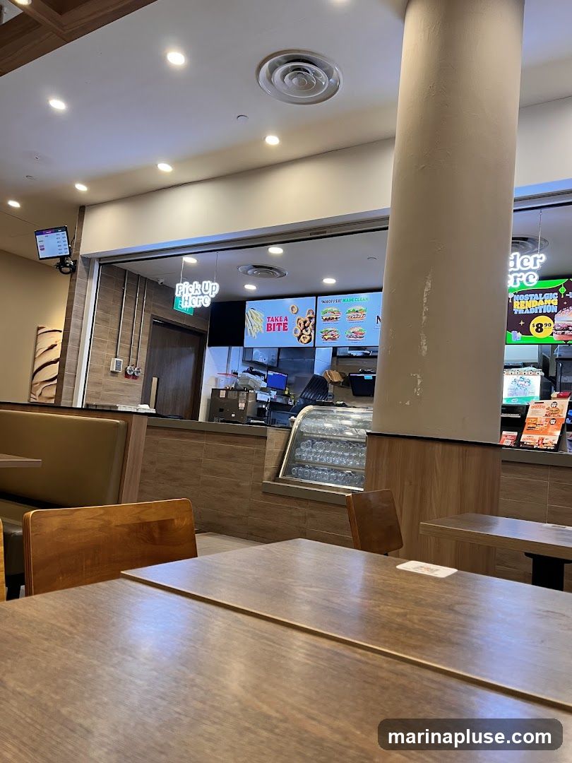 The image shows the interior of a fast-food restaurant. In the foreground, there are wooden tables and chairs. In the background, there is a counter with a menu board displaying food items and prices. A "Pick Up Here" sign is visible above the counter. The overall ambiance is casual and functional.