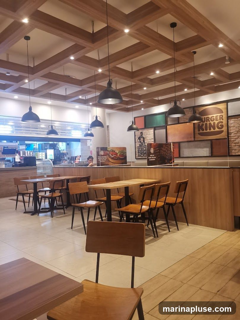 The interior of a Burger King restaurant features a modern design with a coffered ceiling, pendant lights, and wooden accents. Seating includes tables and chairs arranged throughout the dining area. A counter and kitchen area are visible in the background, along with promotional displays featuring burgers and the Burger King logo.