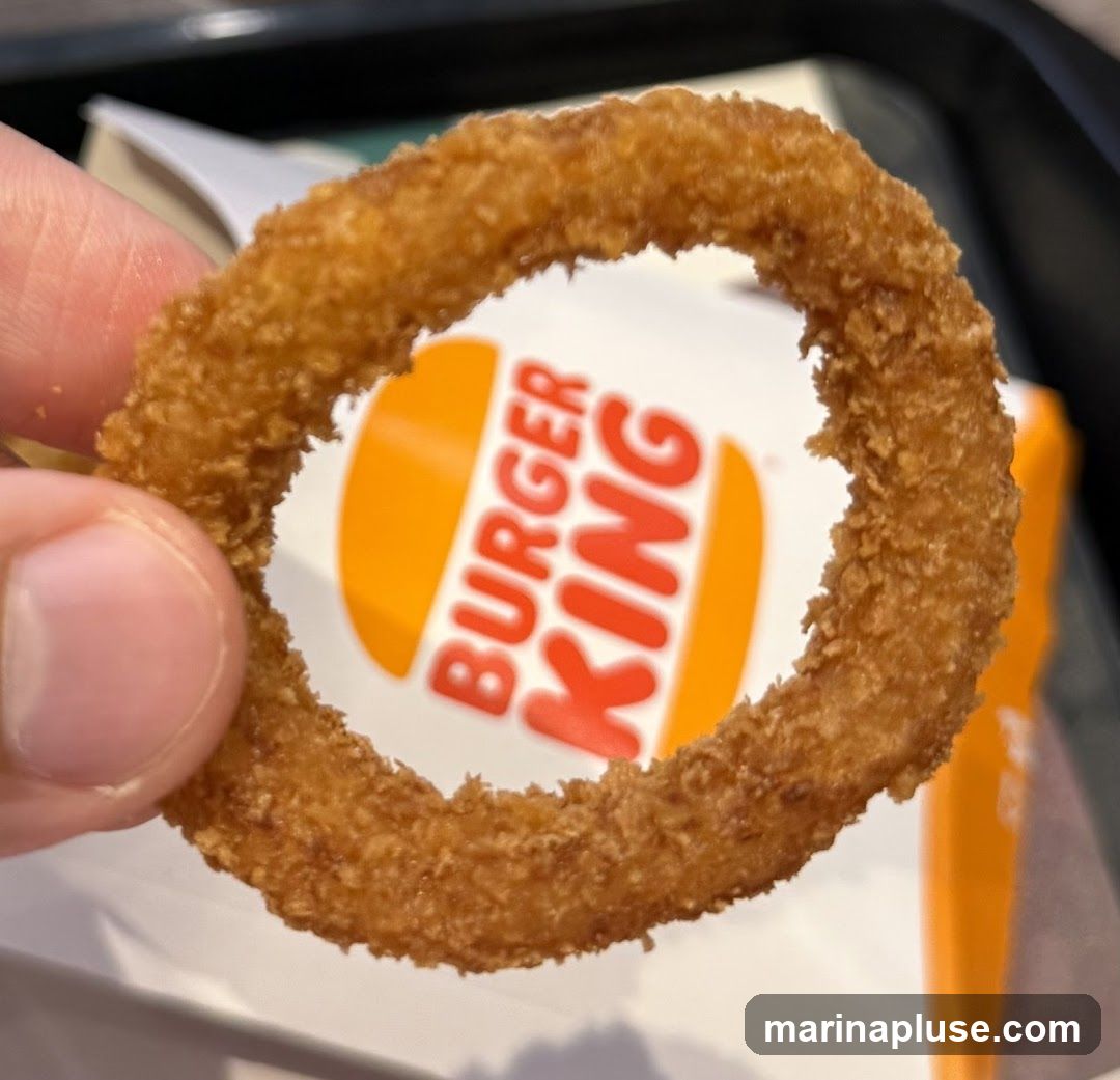 A close-up shot of a hand holding a golden-brown, crispy onion ring. Through the center of the onion ring, the distinctive orange and white Burger King logo is visible on a white background.