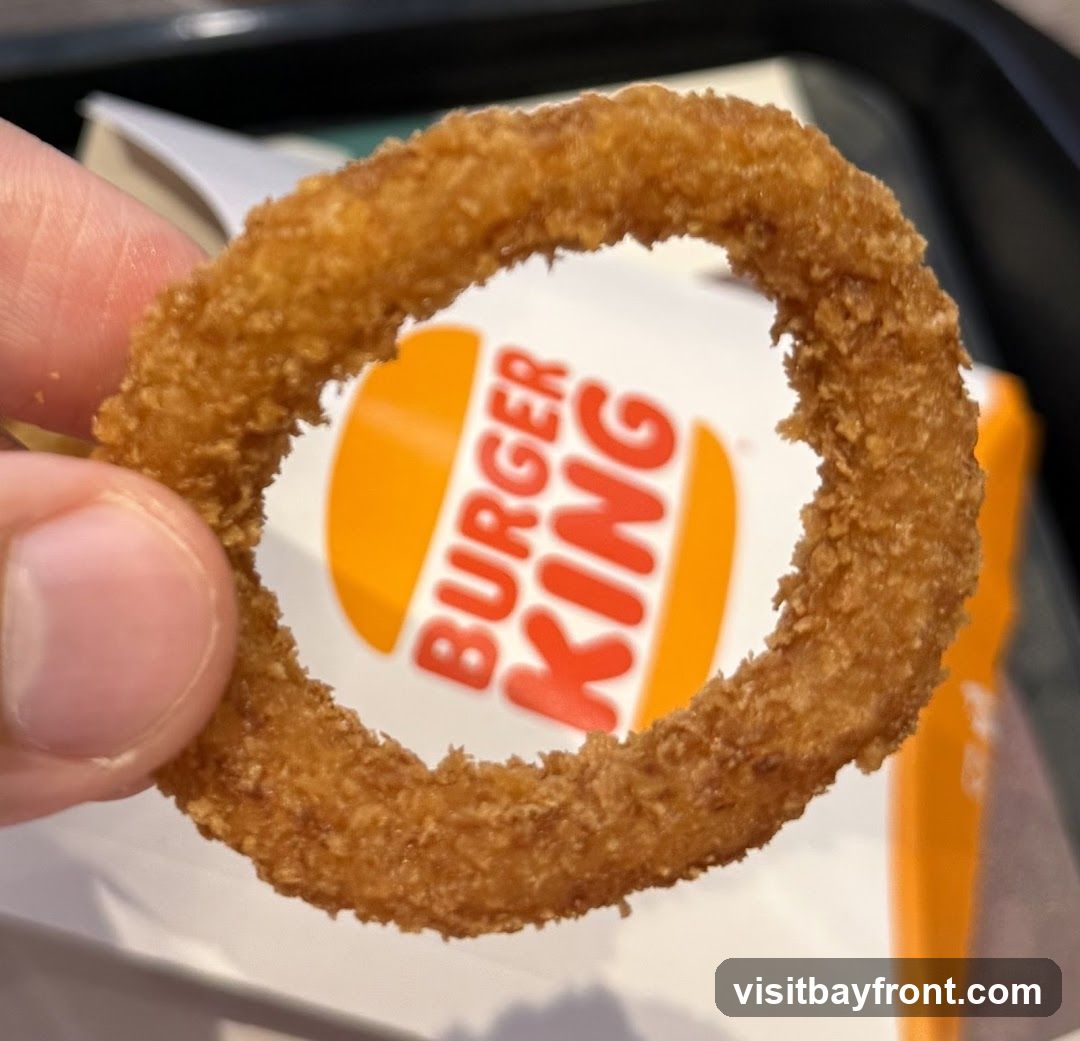 A close-up shot of a hand holding a golden-brown, crispy onion ring. Through the center of the onion ring, the distinctive orange and white Burger King logo is visible on a white background.