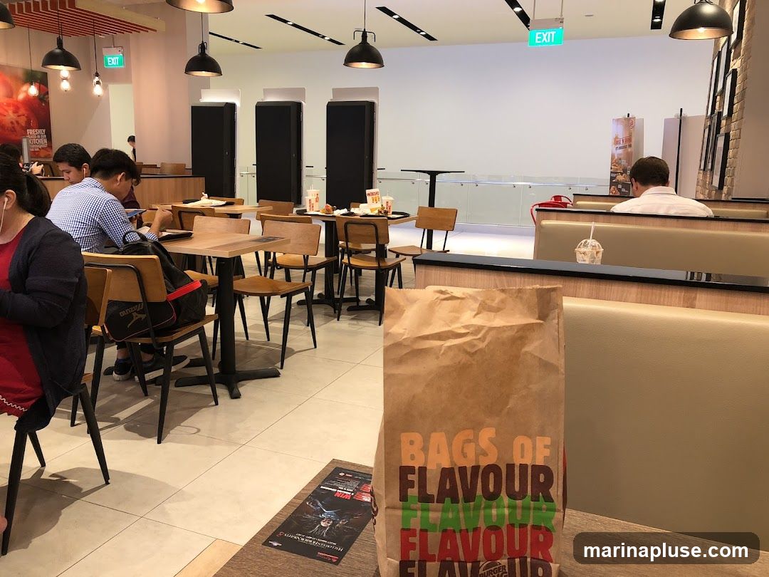 An interior view of a Burger King restaurant with several patrons seated at tables. A brown paper bag with the Burger King logo and the text "BAGS OF FLAVOUR" is prominently displayed in the foreground. The restaurant features light-colored tiled flooring, wooden tables and chairs, and booth seating. Overhead lighting fixtures illuminate the space. An exit sign is visible in the background.