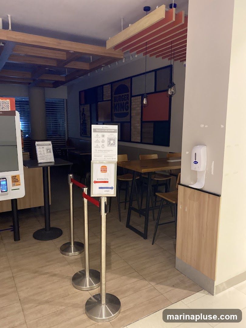 The interior of a Burger King restaurant with a queue management system in place. A sign indicates "Entrance" and "Exit" with arrows. The wall behind the queue features a large Burger King logo and a mosaic of colored panels and brick patterns. Seating areas with tables and chairs are visible in the background. A hand sanitizer dispenser is mounted on a pillar. A self-order kiosk is also present.