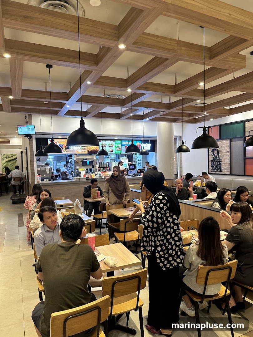 A busy fast food restaurant interior with many people seated at tables. The ceiling has a modern wooden beam design. In the background, a counter area is visible with staff serving customers. Digital menu boards are displayed above the counter.