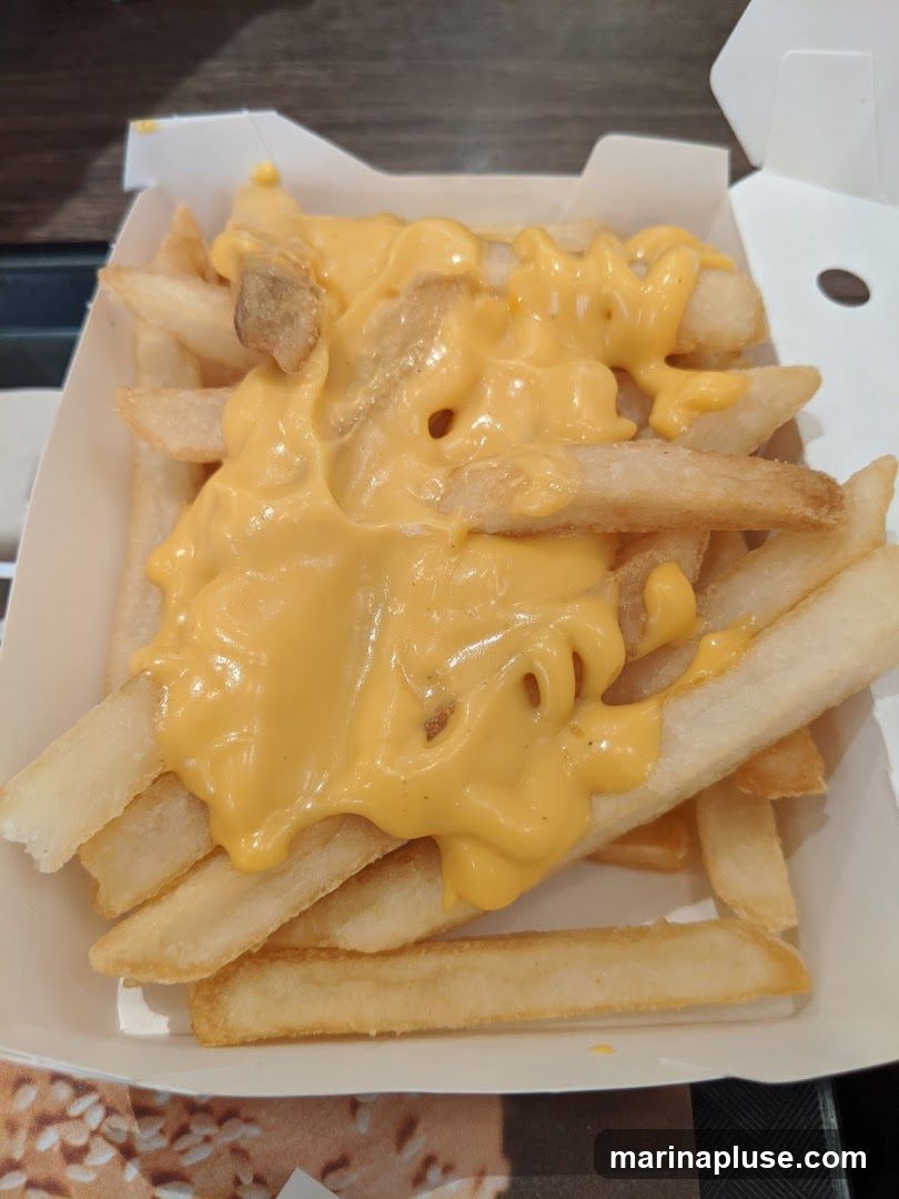 A close-up shot of french fries covered in melted cheese sauce. The fries are golden brown and appear crispy, served in a white cardboard container. The cheese sauce is a vibrant yellow and has a smooth, liquid consistency, drizzled generously over the fries.