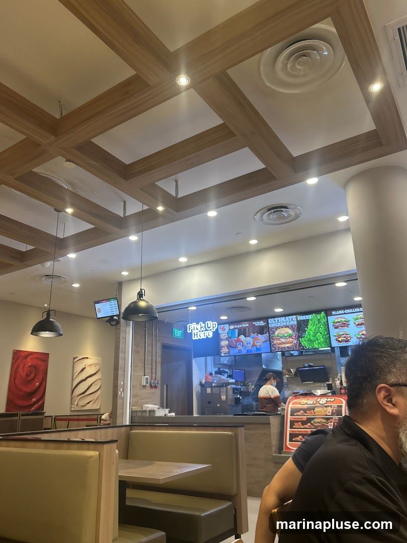 The image shows the interior of a fast-food restaurant with booth seating and a counter area. The ceiling has a modern design with wooden beams and recessed lighting. A digital menu board is visible above the counter, displaying various food items and promotions. A staff member is seen working behind the counter. The overall ambiance suggests a casual dining experience.