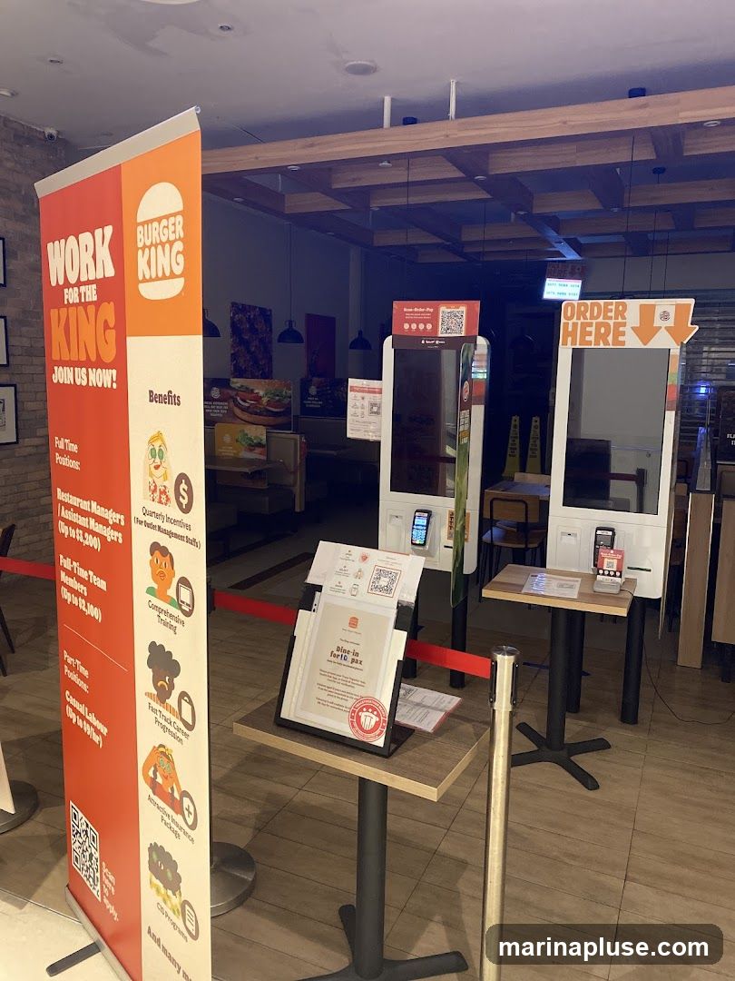 The image shows the interior of a Burger King restaurant. A large vertical banner on the left advertises job openings with the slogan "WORK FOR THE KING JOIN US NOW!". It lists various positions like Restaurant Managers, Assistant Managers, Full-Time Team Members, and Casual Labour, along with benefits such as Comprehensive Training, Fast Track Career Progression, Attractive Insurance Package, and CIS Program. In the background, there are self-order kiosks and tables. A sign on a table reads "Dine-in for 10 pax".