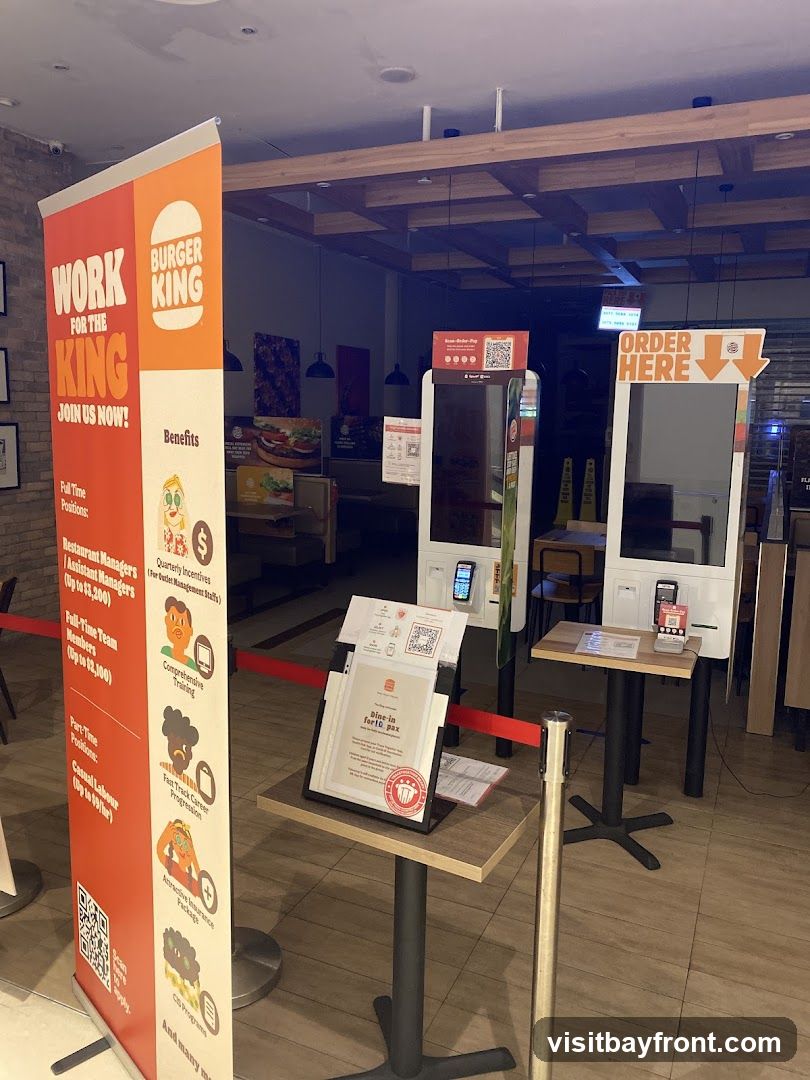 The image shows the interior of a Burger King restaurant. A large vertical banner on the left advertises job openings with the slogan "WORK FOR THE KING JOIN US NOW!". It lists various positions like Restaurant Managers, Assistant Managers, Full-Time Team Members, and Casual Labour, along with benefits such as Comprehensive Training, Fast Track Career Progression, Attractive Insurance Package, and CIS Program. In the background, there are self-order kiosks and tables. A sign on a table reads "Dine-in for 10 pax".