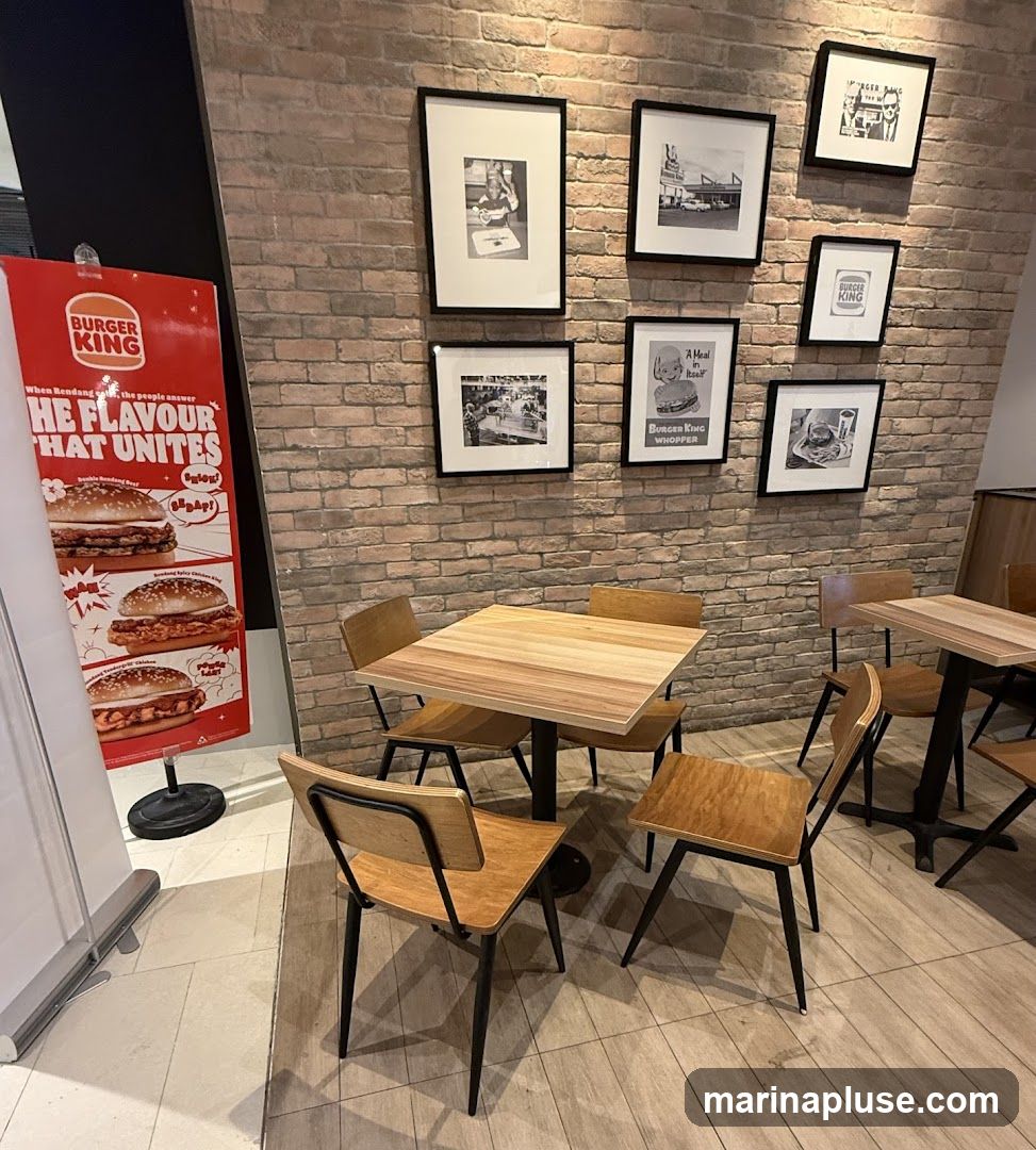 A Burger King restaurant interior with tables and chairs. A promotional poster for Burger King is visible on the left, featuring images of burgers and text about "The Flavour That Unites". The wall is decorated with framed black and white photographs, some of which appear to be historical Burger King advertisements or images.