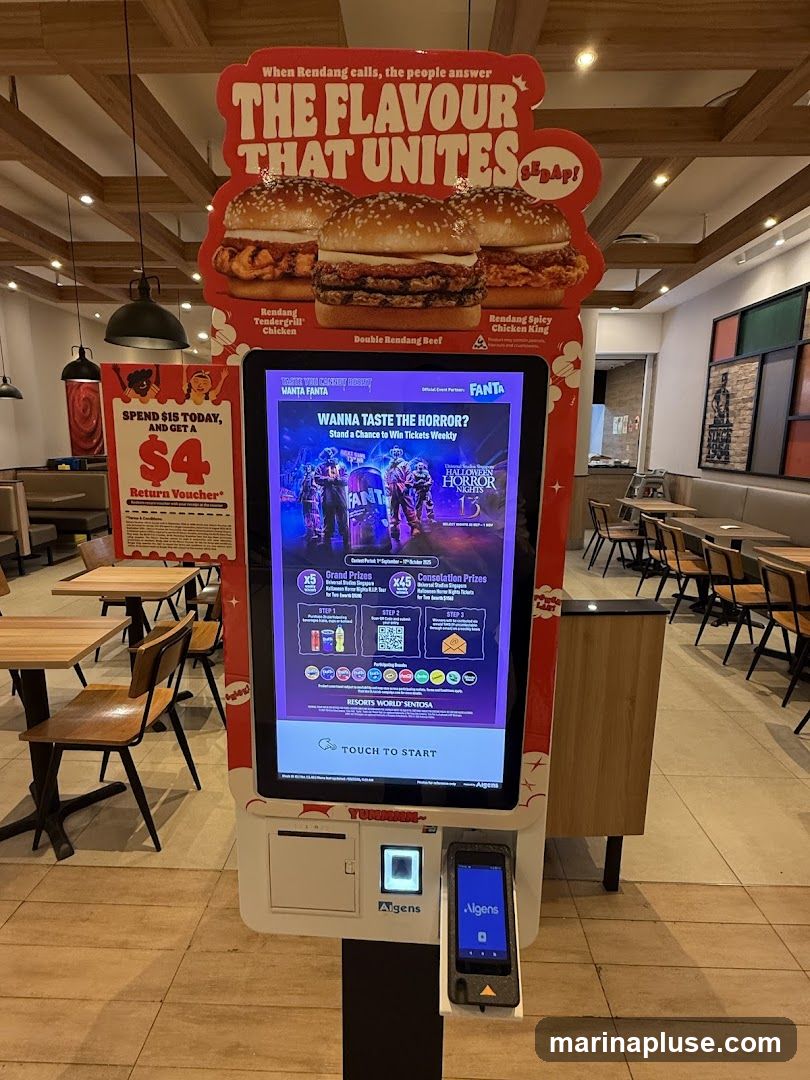 A digital kiosk displays advertisements for burgers and a Halloween event. The top of the kiosk features images of three burgers with text promoting "The Flavour That Unites." Below this, a large screen advertises a chance to win tickets to "Halloween Horror Nights" by purchasing Fanta. A separate sign offers a $4 return voucher with a $15 spend. The kiosk has a payment terminal and a receipt printer. The background shows the interior of a restaurant with tables and chairs.