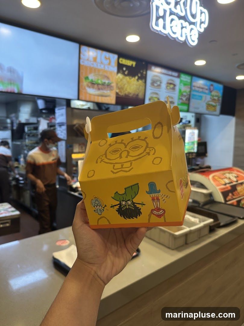 A hand holds a yellow food box decorated with SpongeBob SquarePants characters. In the background, a fast-food restaurant counter is visible with digital menu boards displaying food items like "Spicy Chicken King" and "Crispy Chicken". A "Pick Up Here" sign is illuminated above.