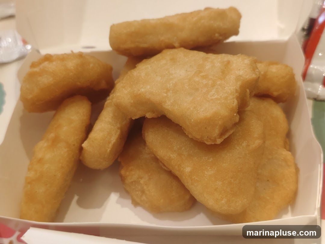 A close-up shot of a white box filled with golden-brown chicken nuggets. Some nuggets are in the shape of traditional nuggets, while others are in fun shapes like a boot and a bell. The nuggets appear to be freshly fried and are piled on top of each other.