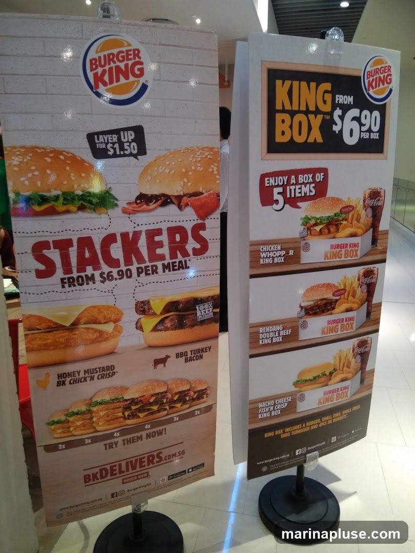 Two promotional stands for Burger King. The left stand advertises "Stackers" meals starting from $6.90, with options to "Layer Up" for $1.50. It showcases Honey Mustard BK Chick'n Crisp and BBQ Turkey Bacon. The right stand promotes "King Box" meals for $6.90, each containing 5 items. Featured King Boxes include Chicken Whopper, Rendang Double Beef, and Nacho Cheese Fish'n Crisp.