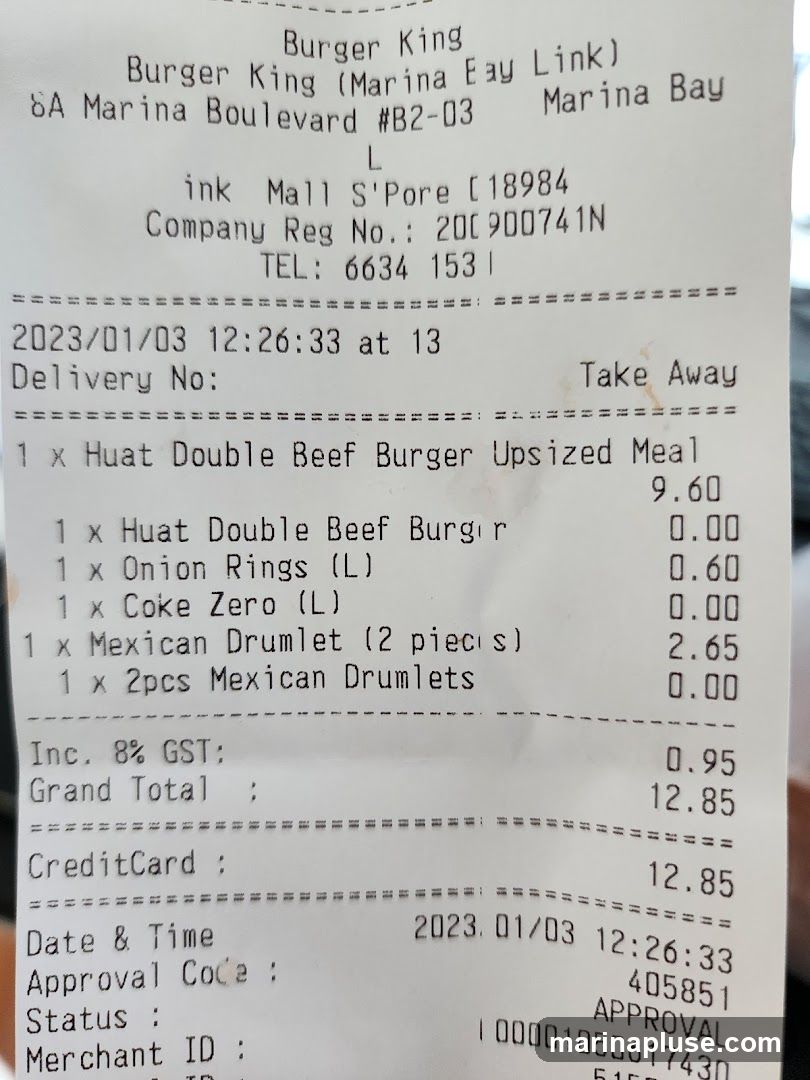 A receipt from Burger King detailing a take-away order. The order includes a Huat Double Beef Burger Upsized Meal, a Huat Double Beef Burger, Onion Rings, Coke Zero, and Mexican Drumlets. The total cost including GST is 12.85.