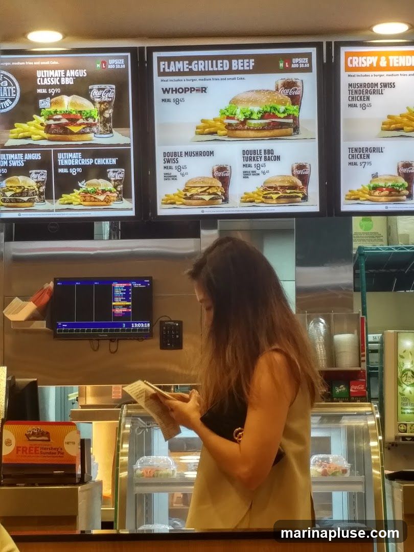 A woman is reading a menu in front of a fast-food counter. Above her, a large menu board displays various burger and chicken sandwich meals with prices. The menu board is divided into sections like "FLAME-GRILLED BEEF" and "CRISPY & TENDER". Below the menu board, a display case shows food items, and a promotional sign for a free Hershey's Sundae Pie is visible.