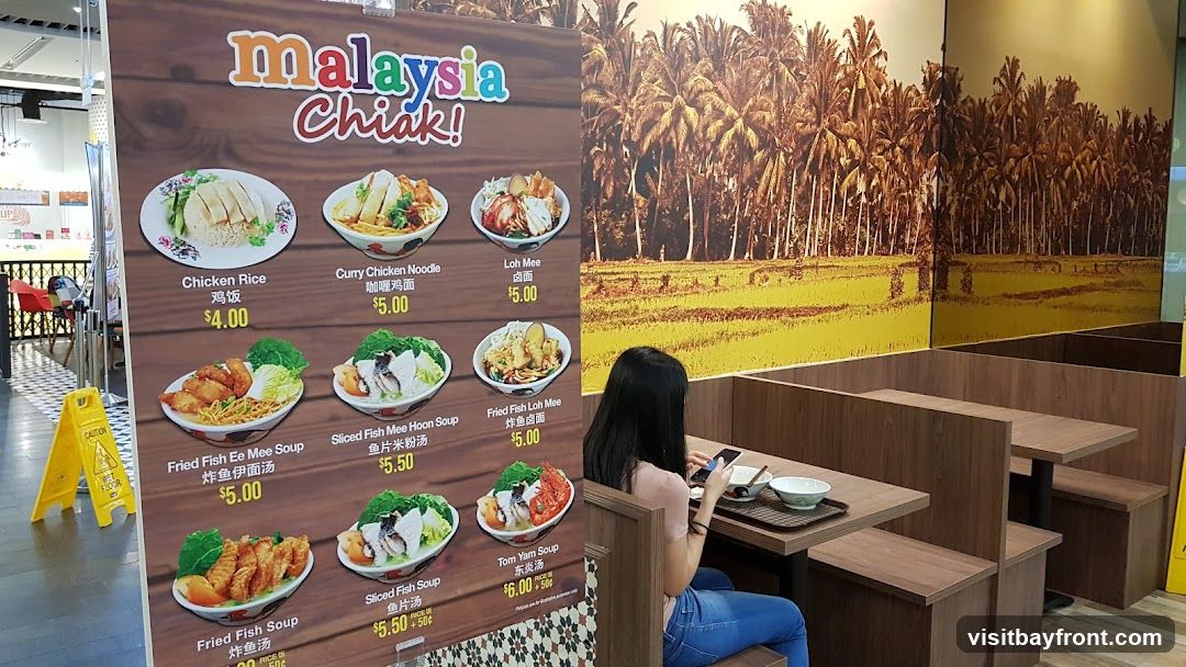 A menu board displays various Malaysian dishes with images and prices. Dishes include Chicken Rice, Curry Chicken Noodle, Loh Mee, Fried Fish Ee Mee Soup, Sliced Fish Mee Hoon Soup, Fried Fish Loh Mee, Fried Fish Soup, Sliced Fish Soup, and Tom Yam Soup. The background features a scenic print of palm trees and a rice field. A woman is seated at a table, looking at her phone.