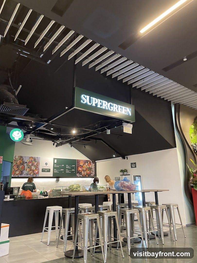 A view of the interior of a Supergreen food establishment, showing a counter with staff preparing food. A large sign with the name "SUPERGREEN" is visible above the counter. There is a communal table with stools in the foreground. A menu board with food images and prices is displayed on the wall.