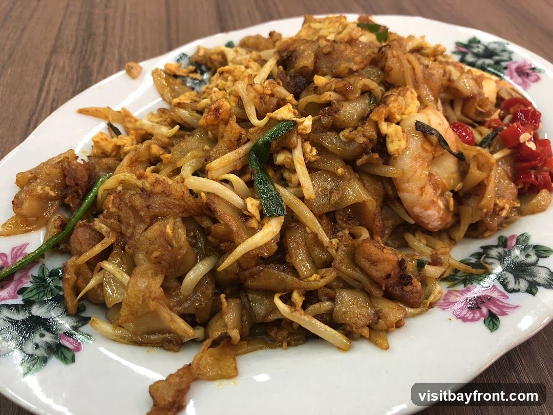 A close-up shot of a plate of Char Kway Teow, a popular stir-fried noodle dish. The dish features flat rice noodles stir-fried with bean sprouts, scrambled eggs, and shrimp. It is garnished with chopped red chilies and green onions. The plate has a floral pattern around the rim.