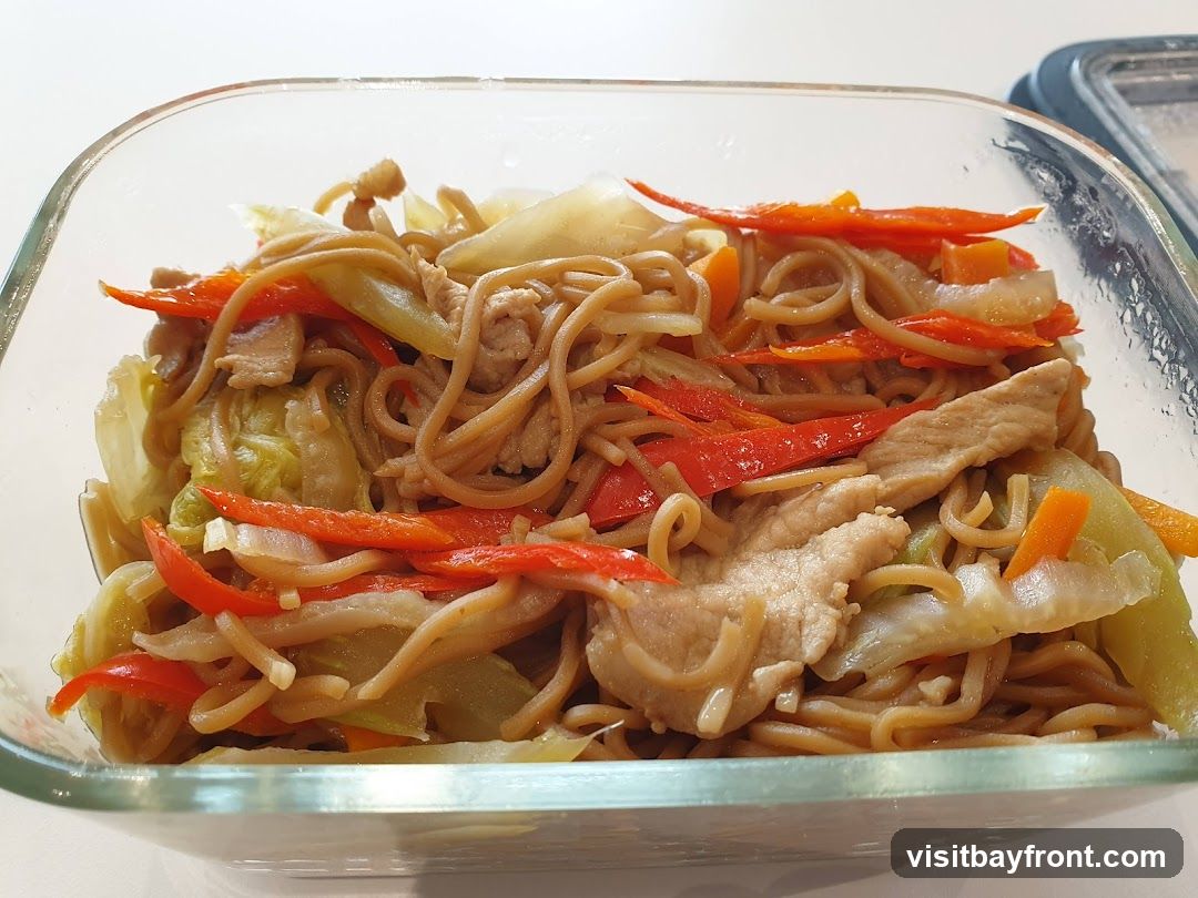 A close-up shot of a glass container filled with stir-fried noodles. The dish contains thin brown noodles mixed with sliced pork, shredded cabbage, julienned carrots, and red bell peppers. The food appears to be seasoned and cooked, ready to be served or stored.
