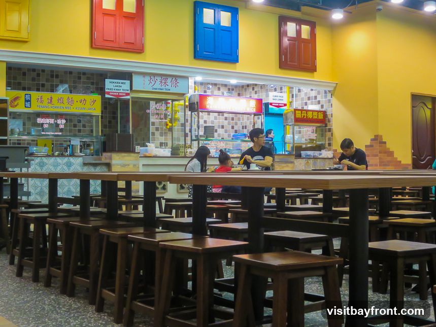 A food court with multiple stalls and seating areas. People are seen eating at tables. The stalls have various signs in Chinese and English advertising food items.
