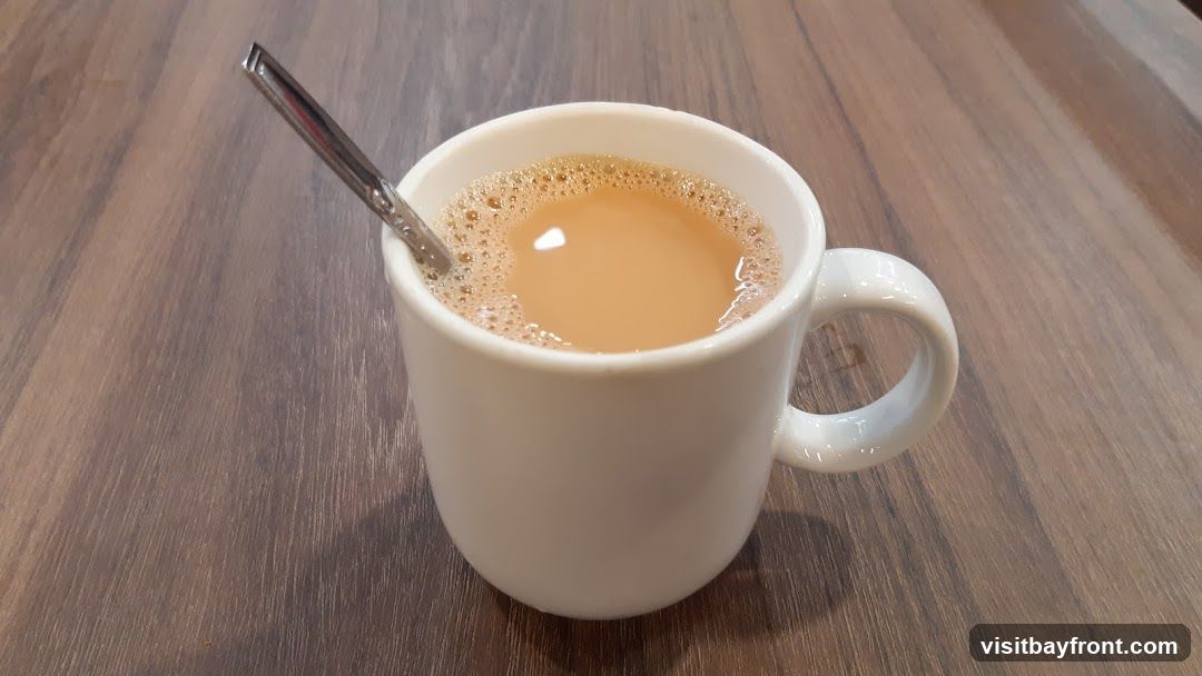 A white mug filled with tea and a spoon resting inside is placed on a wooden table. The tea has a frothy surface.