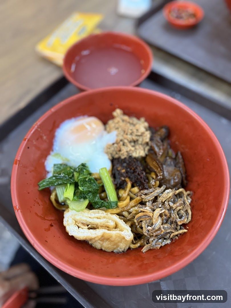 A close-up shot of a bowl of noodles served in a red bowl. The noodles are topped with a soft-boiled egg, minced meat, vegetables, fried anchovies, and a piece of fried tofu. A small bowl of soup is visible in the background.