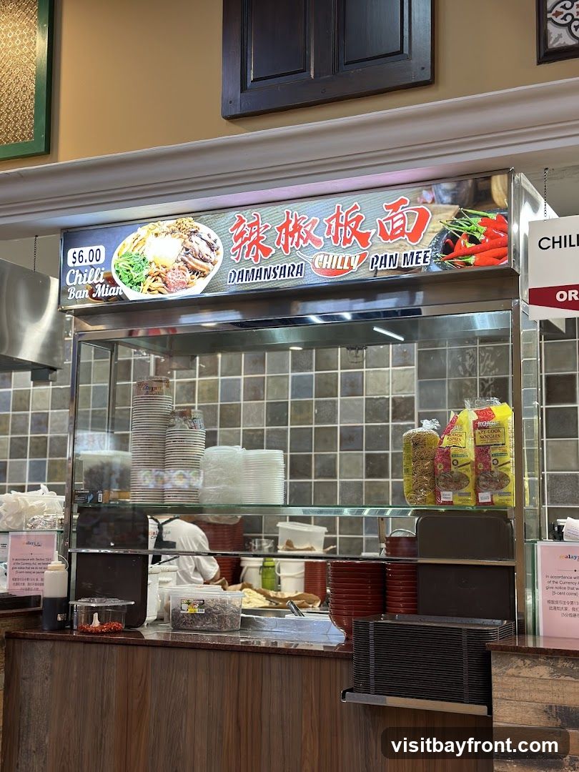 A food stall display featuring a large illuminated sign with a picture of a noodle dish and chili peppers. The sign reads "$6.00 Chilli Ban Mian" and "DAMANSARA CHILLY PAN MEE" in English and Chinese characters. Below the sign, shelves are stocked with stacks of bowls, plates, and packets of noodles. The counter area shows various ingredients and condiments.
