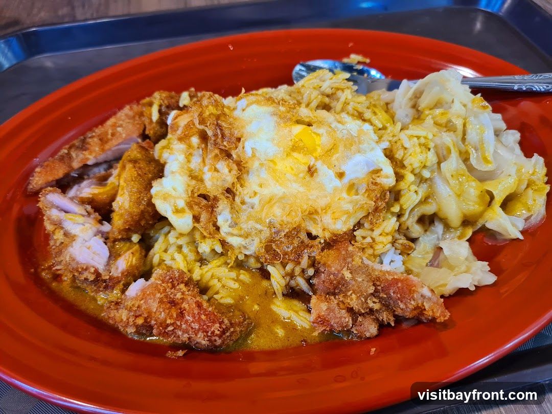 A close-up shot of a plate of food, featuring rice topped with fried chicken pieces, a sunny-side-up egg, and a side of braised cabbage. The dish is served on a red oval plate, which sits on a dark tray. A spoon is visible on the right side of the plate.