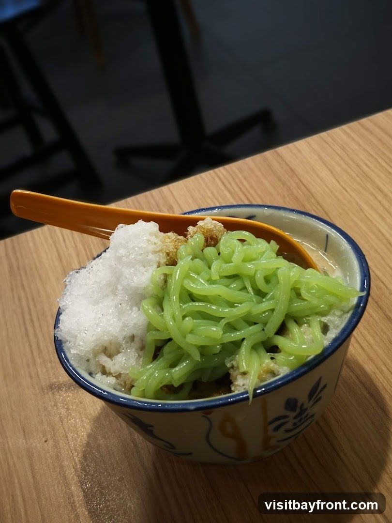 A bowl of cendol, a traditional dessert, is presented on a wooden table. The dessert features green jelly noodles, shaved ice, and brown sugar syrup, topped with a spoon. The bowl has a blue and white decorative pattern. In the background, blurred dark shapes suggest tables and chairs, indicating an indoor setting.