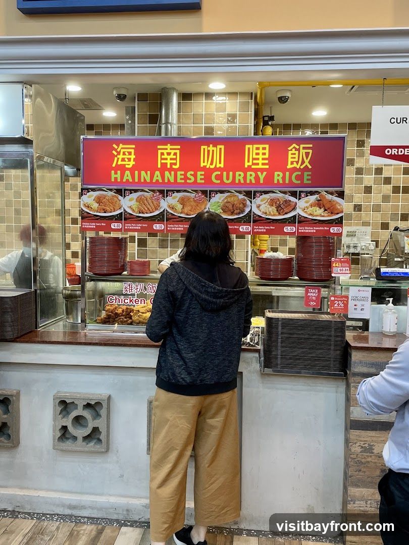 A person is standing in front of a food stall that serves Hainanese Curry Rice. The stall has a large red sign with Chinese characters and the English text "HAINANESE CURRY RICE". Below the sign are pictures of various curry rice dishes with prices. The stall displays fried chicken and other food items behind a glass counter.