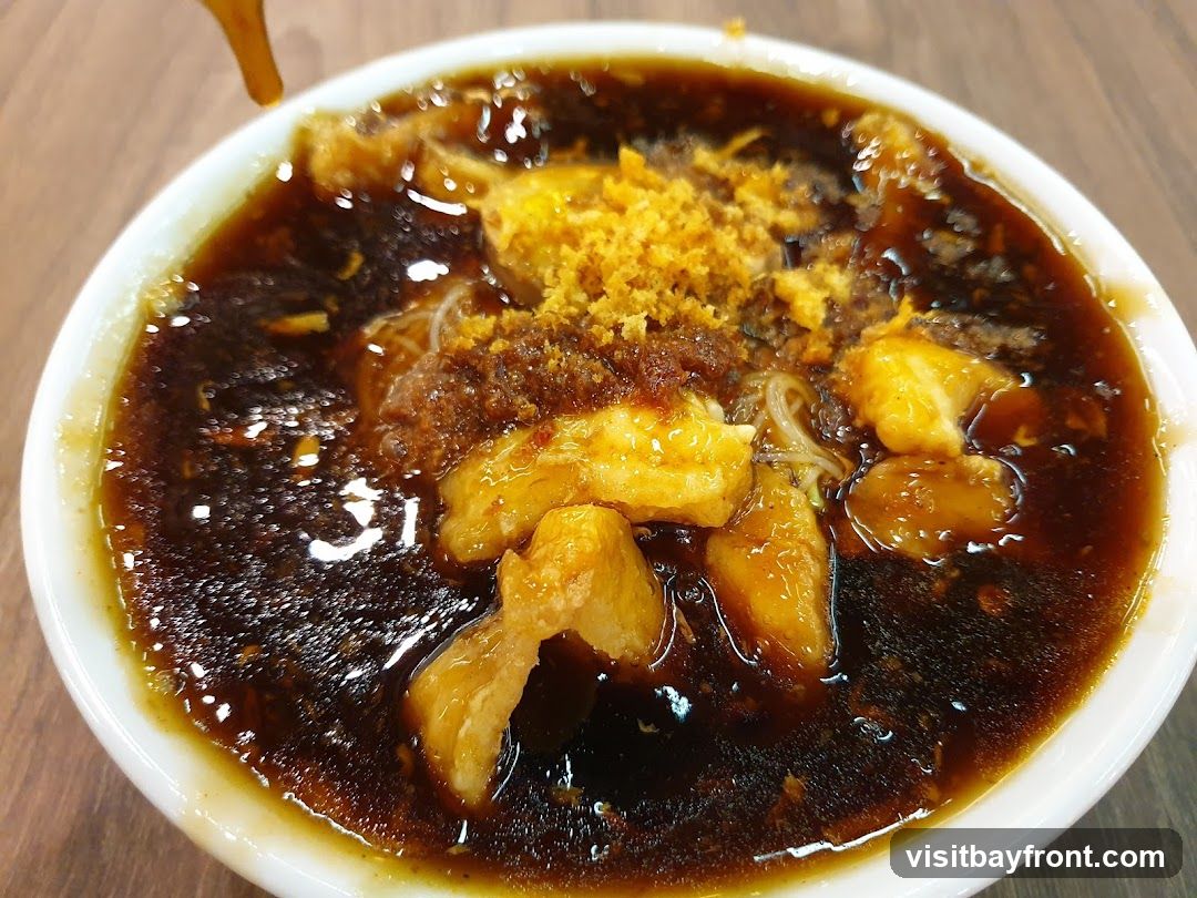 A close-up shot of a white bowl filled with dark broth, noodles, and various toppings. The toppings include pieces of fried dough, a sprinkle of crispy brown bits, and what appears to be minced garlic or fried shallots. A thin stream of dark liquid is being poured into the bowl from the top left. The bowl is placed on a wooden surface.