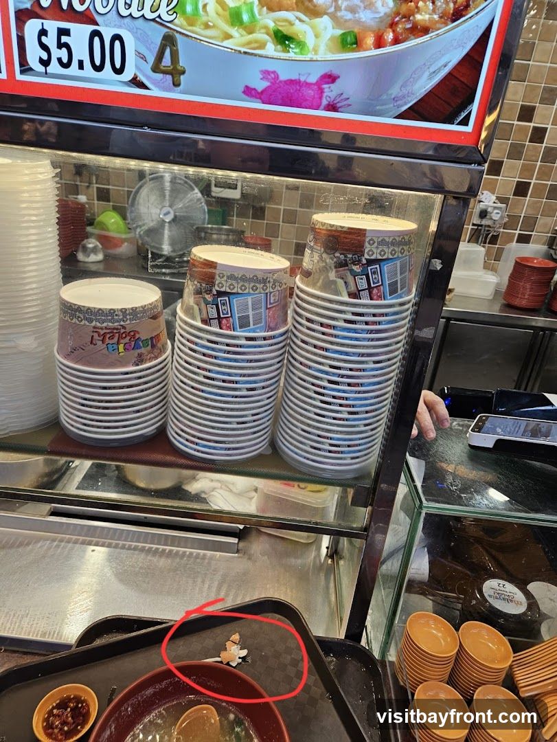 A close-up view of a food stall counter, showing stacks of disposable cups and bowls. Above, a sign displays a bowl of noodles with the price '$5.00' and the number '4'. A tray with a bowl of soup and a small dish of dipping sauce is visible in the foreground. A small pile of what appears to be broken eggshells is circled on the tray.