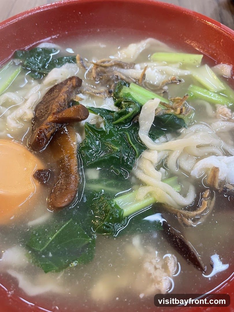 A close-up shot of a bowl of noodle soup. The soup contains wide, flat noodles, leafy green vegetables, sliced mushrooms, and what appear to be small dried fish. The bowl is red and has a textured rim.