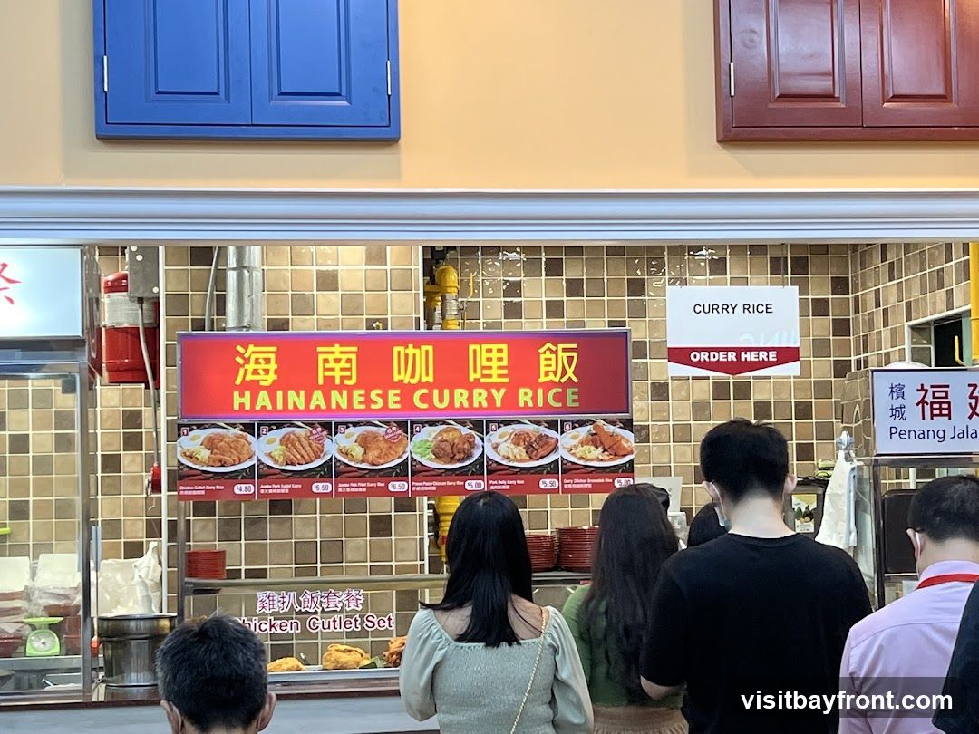 A food stall with a prominent sign for Hainanese Curry Rice. Several dishes are displayed with prices. People are queuing to order.