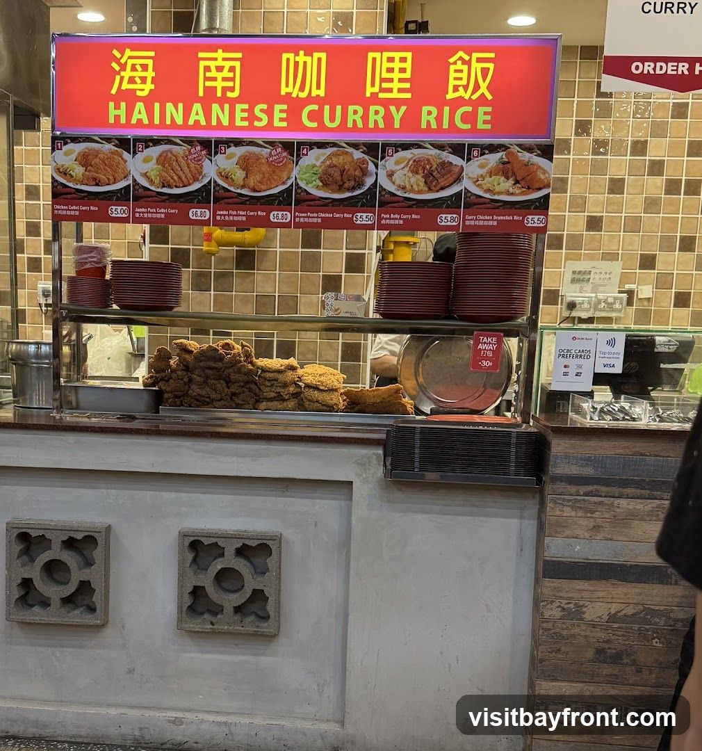 A food stall with a prominent sign displaying "Hainanese Curry Rice" in both Chinese and English. Below the sign, there are pictures and prices for various curry rice dishes. Piles of fried food are visible, along with stacks of plates and a "Take Away" sign. Payment options, including credit cards and contactless payment, are displayed.