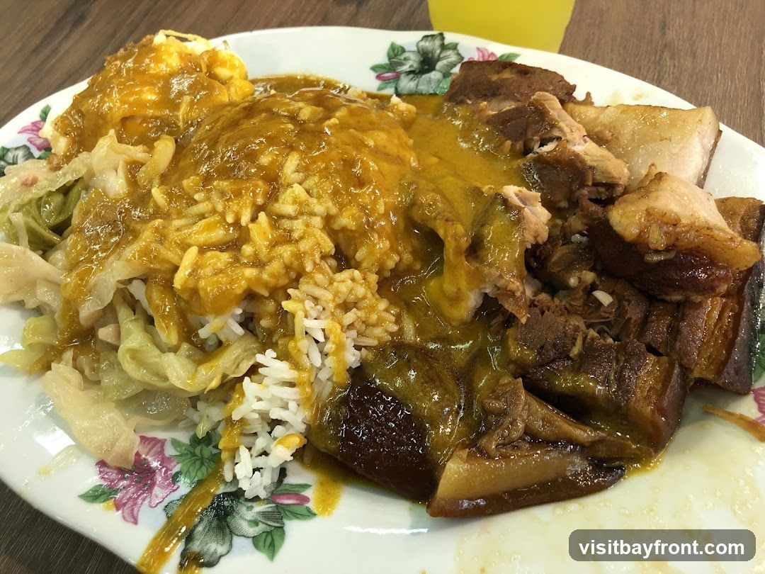A close-up shot of a plate of curry rice. The dish features white rice topped with a generous amount of yellow curry sauce. Alongside the rice are braised pork pieces, some with visible fat and skin, and a portion of stewed cabbage. A fried egg with a runny yolk is also visible. The plate has a floral pattern around the rim. A glass of yellow liquid, possibly a drink, is partially visible in the upper right corner.