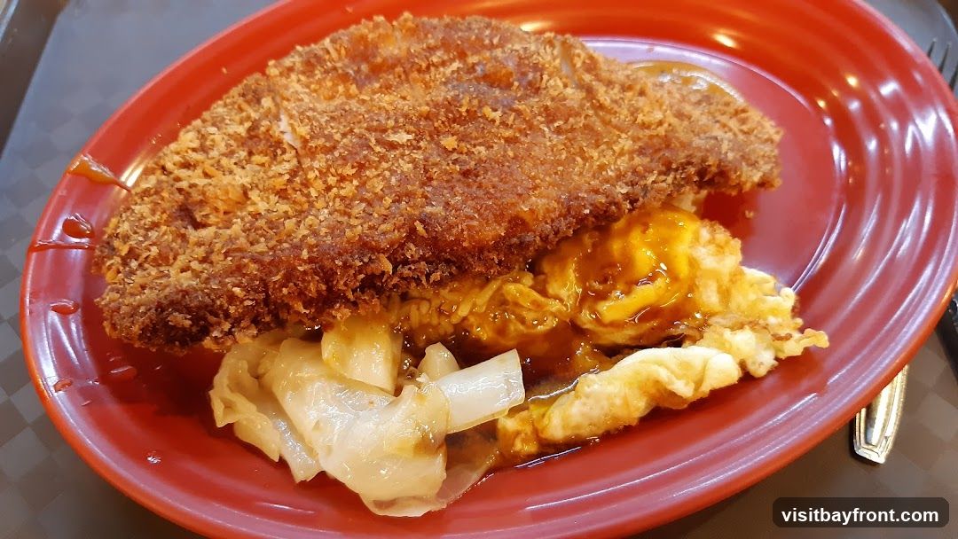 A close-up shot of a meal served on a red plate. The dish features a large, breaded and fried chicken cutlet placed over a bed of rice. Scrambled eggs, coated in a savory sauce, are visible beneath the cutlet, along with pieces of braised cabbage. The plate has a few drips of sauce on its rim.
