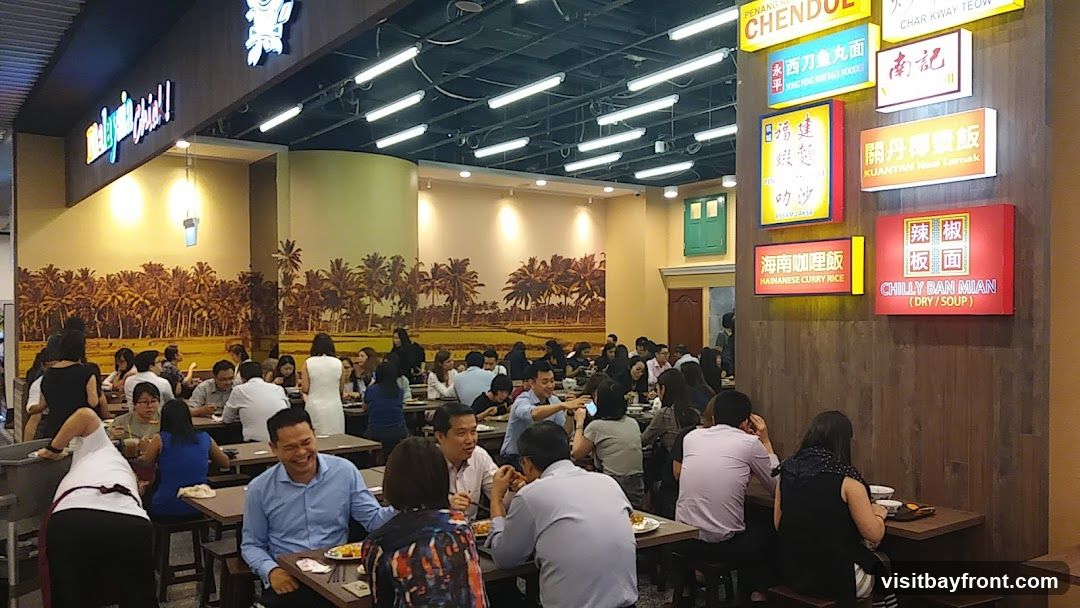 A busy Malaysian food restaurant with many patrons seated at tables. The walls are decorated with a mural of a tropical landscape and several illuminated signs displaying menu items in both Chinese and English. The signs advertise dishes such as Penang Chendol, Char Kway Teow, Yong Peng Fish Ball Noodles, Kuantan Nasi Lemak, Hainanese Curry Rice, and Chilly Ban Mian.