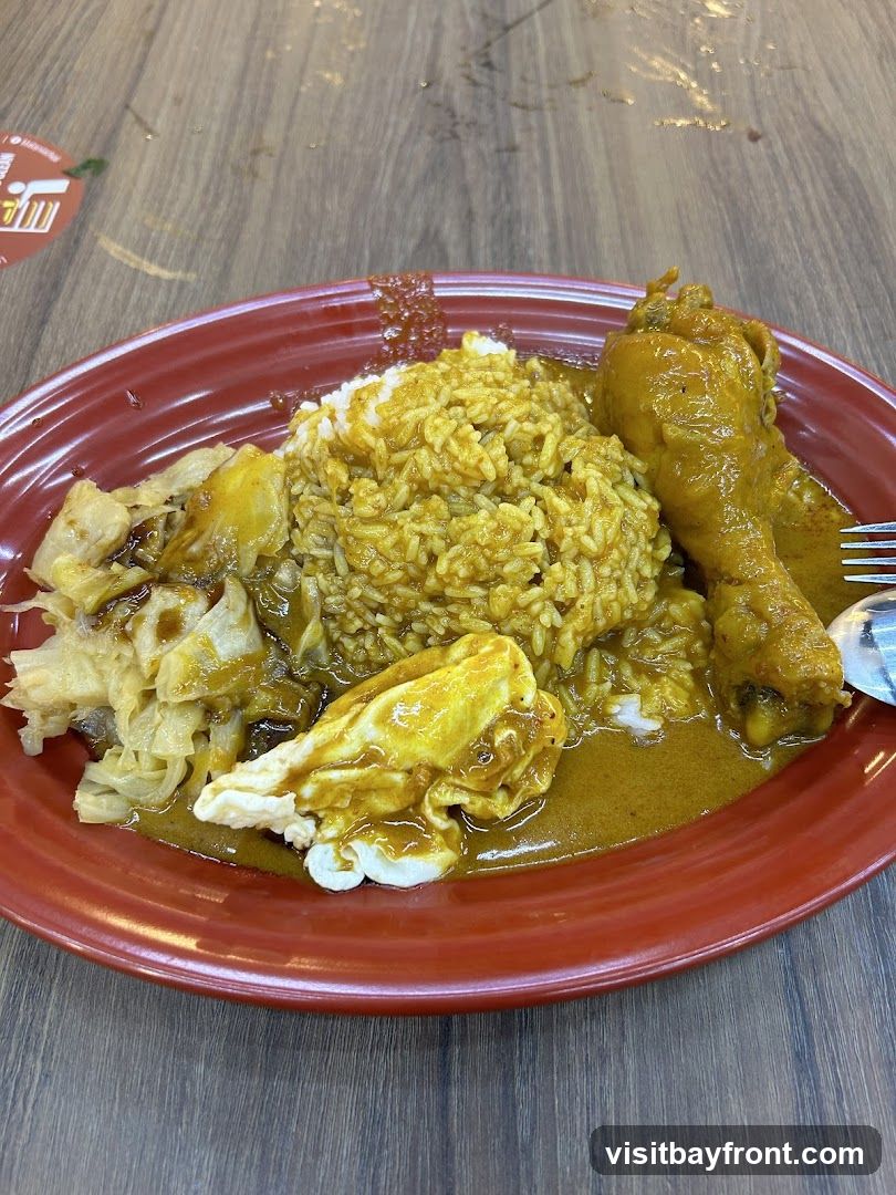 A plate of rice with curry chicken and other side dishes. The rice is topped with a yellow curry sauce. A chicken drumstick is placed on the side of the rice, also covered in curry sauce. There are also some braised vegetables and a fried egg with curry sauce. The plate is red and sits on a wooden table. A fork and spoon are visible on the right side of the plate. A small red sticker with white text and a graphic is on the table to the left of the plate.