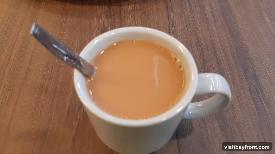 A close-up shot of a white mug filled with a light brown beverage, likely tea with milk. A silver spoon is resting inside the mug. The mug is placed on a wooden surface.