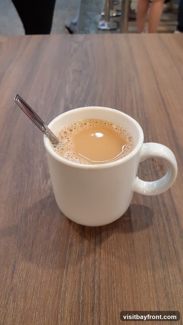 A white mug filled with tea and a spoon rests on a wooden table. In the background, blurred figures of people can be seen.