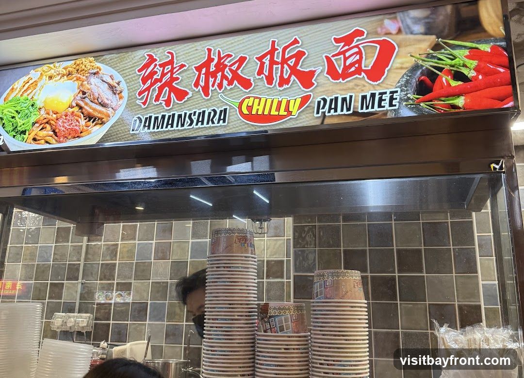 A brightly lit sign displays a bowl of pan mee, chilies, and the text "DAMANSARA CHILLY PAN MEE" in English and Chinese. Below the sign, stacks of paper cups and plates are visible, along with a person wearing a mask in the background. The wall behind is tiled.