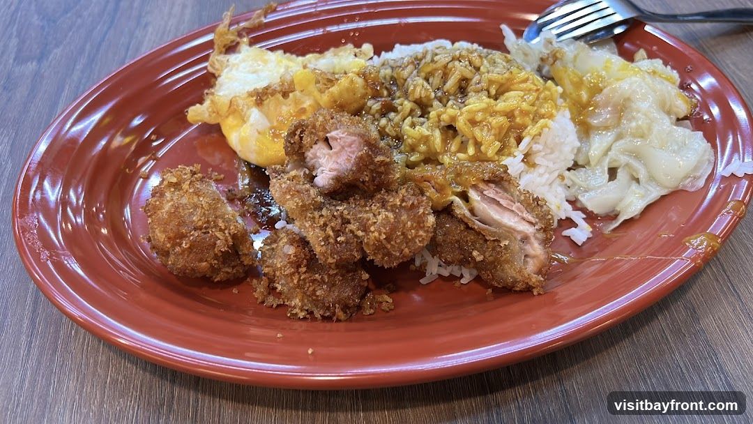 A close-up shot of a plate of food, featuring white rice topped with a yellow curry sauce. To the left of the rice are several pieces of crispy fried chicken. A fried egg with a runny yolk is also visible on the plate, along with some steamed dumplings or wontons. A fork and knife are partially visible at the top right. The plate is a reddish-brown color and rests on a wooden surface.
