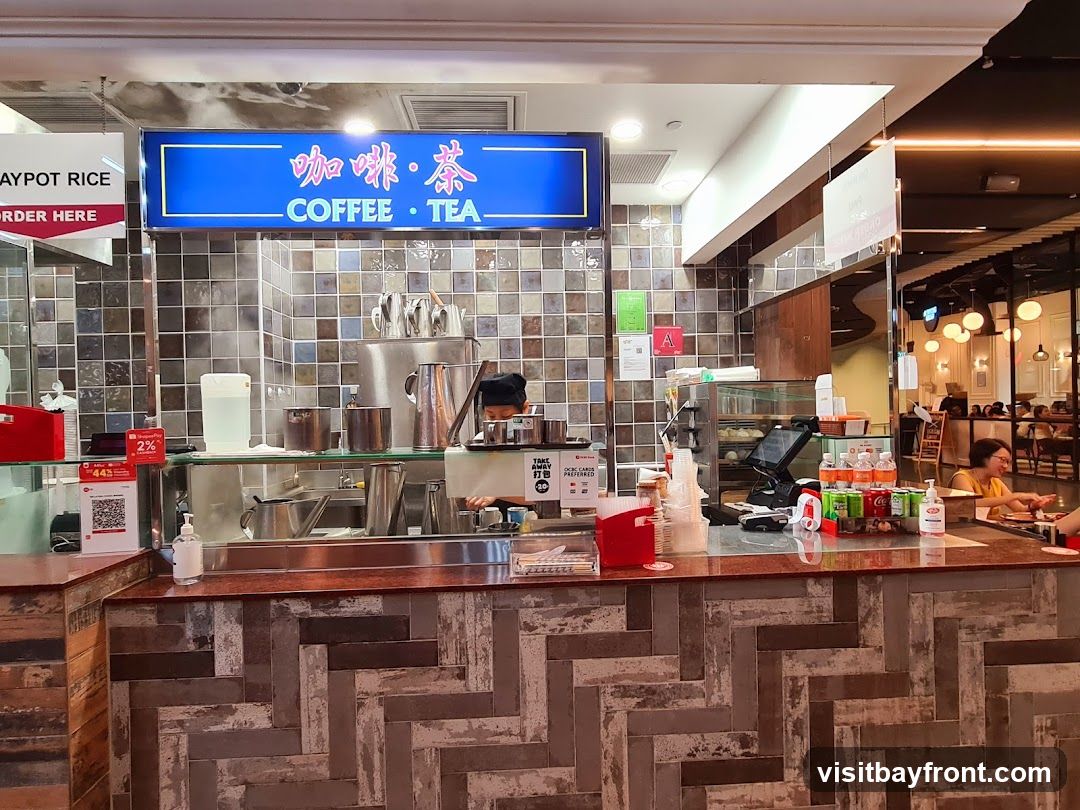 A coffee and tea stall with a blue sign that says "COFFEE • TEA" in English and Chinese characters. There is a person behind the counter preparing drinks. Various beverages and supplies are visible on the counter and shelves. In the background, there is a seating area with other patrons.