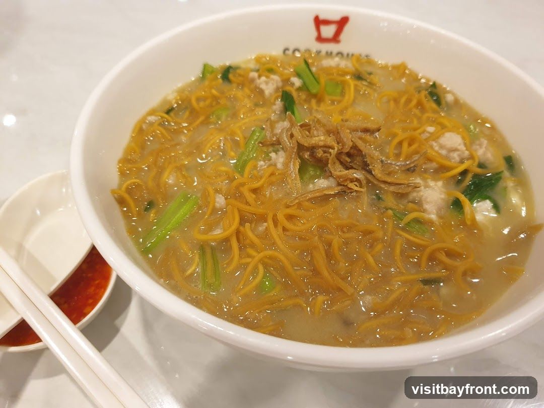 A close-up shot of a bowl of noodles with minced meat, green vegetables, and fried anchovies. The noodles are thin and golden brown, submerged in a light broth. A small dish of red chili sauce and a pair of white chopsticks are placed to the side. The bowl has a logo that reads "COOKHOUSE" with a stylized graphic above it.