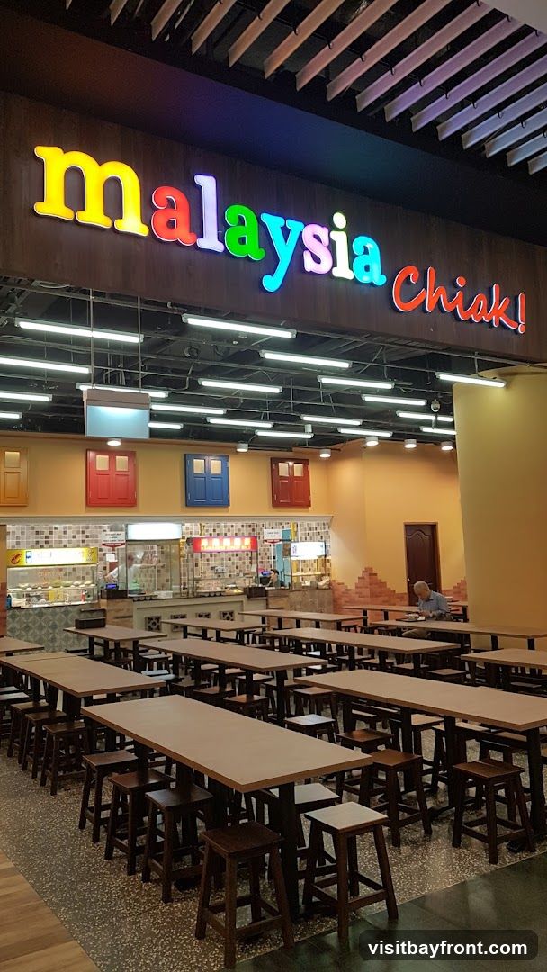 A food court with long tables and stools, featuring a prominent sign that reads "malaysia Chiak!". The background shows various food stalls with colorful decorations, including red, blue, and yellow doors above the stalls. A lone man is seated at one of the tables in the distance.