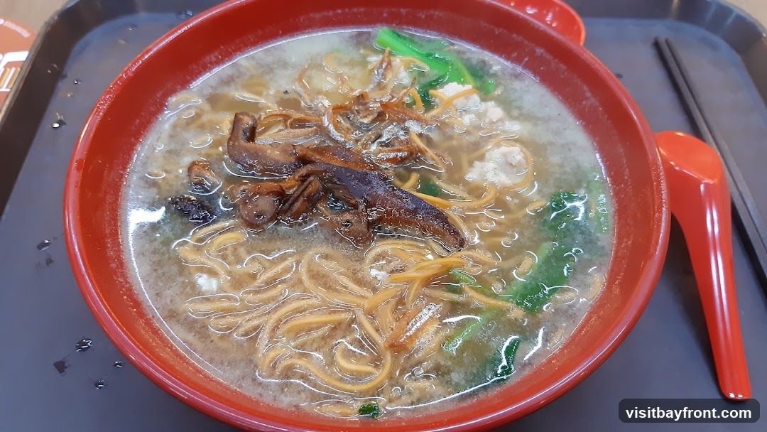 A close-up shot of a bowl of noodle soup. The soup contains thin yellow noodles, minced meat, green vegetables, and pieces of mushroom and dried anchovies. A red spoon and chopsticks are visible on the side.