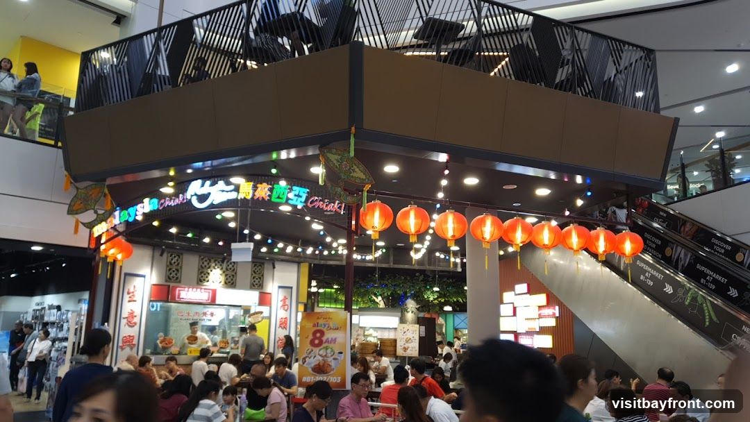 A bustling food court with a Malaysian theme, decorated with red lanterns and kites. People are seated at tables, eating and socializing. An escalator is visible on the right side of the image, with signage for a supermarket.